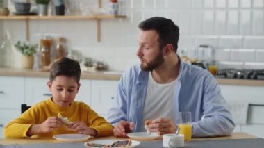 Çocuk mutfakta oturmuş peynirli sandviç yiyor ve babasıyla konuşuyor. Bir adam öğle yemeği sırasında oğluyla iletişim kurar. Yüksek kalite 4k görüntü