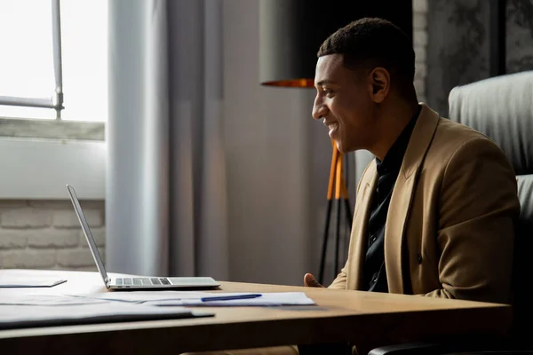 Side view of a black man in a business suit who is sitting in front of a laptop. Smiling businessman sitting at the table and looking at the computer screen. High quality photo