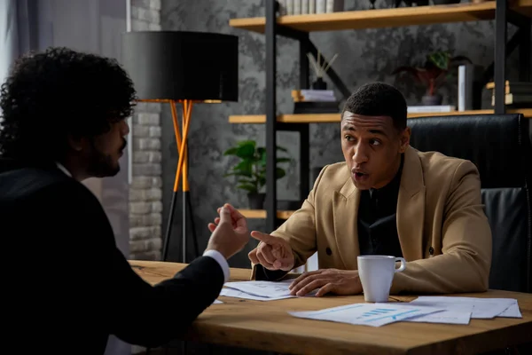 茶色のジャケットを着た黒人男性実業家が彼の労働者を叱った スーツを着た2人の男がオフィスのテーブルに座って何かについて議論している 高品質の写真 — ストック写真