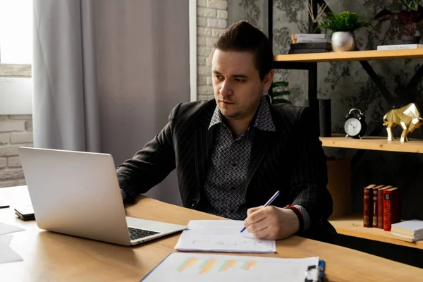 オフィスに座っている間 ビジネススーツの若者がノートパソコンで働いている 黒いジャケットの男がノートパソコンの前に座り 書類を記入する 高品質の写真 — ストック写真