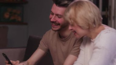 Sarışın, yetişkin oğluyla oturma odasında oturuyor. Ona telefonda bir şey gösteriyor. Bir adam ve annesi kanepede oturmuş komik videolar izlerken gülüyorlar.