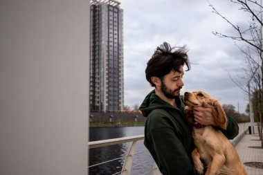 Nehrin kenarında duran sakallı bir adam kollarında bir spanyel köpeği tutuyor ve boynunu çiziyor. Yeşil ceketli bir adam parkta sabah yürüyüşü sırasında köpeğine sarılır. Yüksek kalite fotoğraf