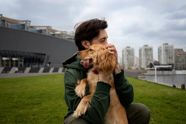 A man in a green jacket hugs his spaniel dog. A man hugs his dog while sitting on the grass against the backdrop of a cloudy city. High quality photo