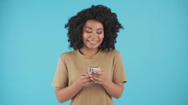 Güzel siyah bir kadın mavi arka planda duruyor ve akıllı telefon kullanıyor. Akıllı telefonuyla sosyal medyada gezinirken kıvırcık saçlı gülümseyen genç bir kadın. Yüksek kalite 4k görüntü