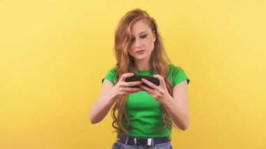 Kızıl saçlı genç bir kadın turuncu bir duvarın karşısında duruyor ve telefonda oynuyor. Yeşil tişörtlü kızıl saçlı bir kadın mobil bir oyunda zor bir aşamadan geçtikten sonra iç çekiyor. Yüksek kalite 4k