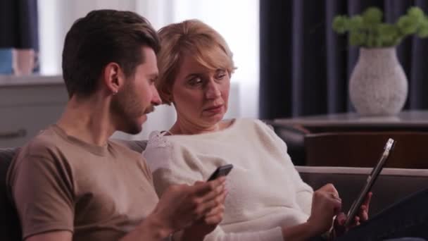 大人の女性がデジタルタブレットを手にソファに座り 隣に座っている息子と話をする 大人の男と彼の母親は リビングルームに座って デジタルデバイスを使用しています — ストック動画