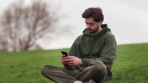 Man Sitter Gräset Och Bläddrar Sociala Nätverk Sin Smartphone Brunett — Stockvideo