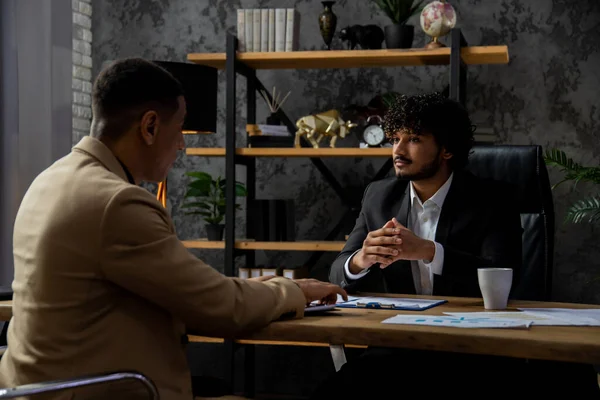 オフィスのテーブルに座っている間 正式なスーツの若者たちはビジネス会話をしています インドのビジネスマンは 彼のビジネスパートナーと議論しています新しいスタートアップの作成 — ストック写真