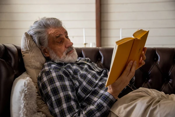 stock image Old man in checkered shirt is lying on sofa and reading book in living room in his house. High quality photo