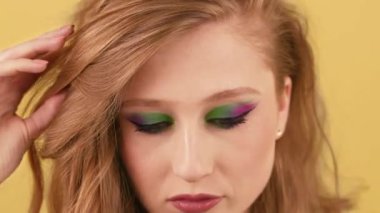 Çekici bayan saçını düzeltip kameraya bakıyor. Göz kapaklarında çok renkli gölgeler olan genç bir kadın turuncu arka planda poz veriyor. Yüksek kalite 4k görüntü