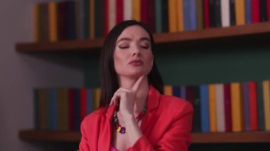 Kırmızı ceketli düşünceli esmer kadın kitaplığın yanında dururken bir şey düşünüyor. Yüksek kalite 4k görüntü