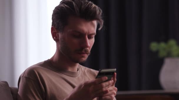 一个皱着眉头 穿着棕色T恤的年轻人坐在沙发上 手里拿着手机 一个满嘴胡茬的年轻人正在他的智能手机上输入一个信息 高质量的4K镜头 — 图库视频影像