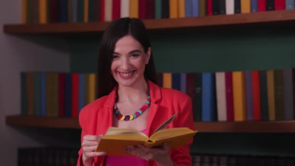 赤いジャケットを着た興奮したブルネットの女性は本を読んで本棚の近くで微笑んだ 本を持っている赤いジャケットの幸せな女性 高品質4K映像 — ストック動画