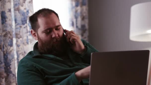 一个满脸胡须的男人坐在笔记本电脑前 在讲电话 一名在家工作的男性自由职业者向雇主报告所做的工作 高质量的4K镜头 — 图库视频影像