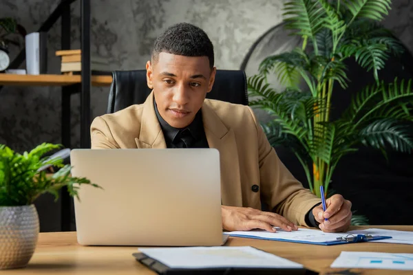 若い黒人男性がオフィスに座って ノートパソコンの画面を注意深く見ています 男性実業家は書類に署名して自宅に座っている 高品質の写真 — ストック写真