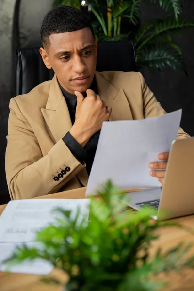 ジャケットを着たアフリカ系アメリカ人男性がオフィスのテーブルに座り 書類を注意深く勉強しています 金融会社の取締役は損益計算書を研究する 高品質の写真 — ストック写真