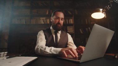 Yelek giymiş sakallı bir adam dizüstü bilgisayarda yazıyor. Erkek bir işadamı evindeki bir masada oturuyor ve bir bilgisayarda çalışıyor. Yüksek kalite 4k görüntü