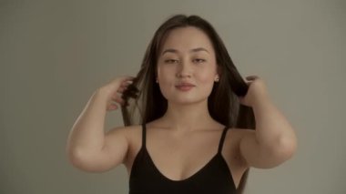 Genç, uzun saçlı bir kadın kameranın önünde duruyor ve saçını düzeltiyor. Gri arka planda duran genç bir kadın parmaklarıyla nazikçe yüzünü okşar. Yüksek kalite 4k görüntü