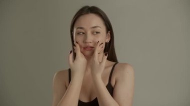 Gri arka planda kameraya poz veren uzun saçlı hoş bir genç kadın. Güzel bir Asyalı kadın kameranın önünde duruyor ve nazikçe yüzünü okşuyor. Yüksek kalite 4k görüntü