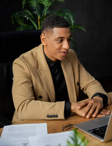 正式なスーツを着た黒人がオフィスに座り 笑顔でノートパソコンで何かを見ている アフリカ系アメリカ人ビジネスマンはビデオリンクを介してビジネスパートナーと通信します 高品質の写真 — ストック写真