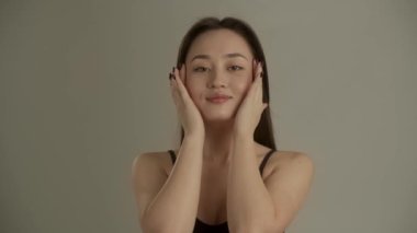Gri bir duvarın arkasında dikilirken kameranın önünde poz veren gülümseyen esmer kadın. Güzel bir Asyalı kadın kameranın önünde duruyor ve nazikçe yüzüne masaj yapıyor.