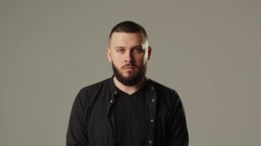 Gri arka planda kameraya poz veren genç sakallı adam. Siyah kazaklı bir adam gri bir duvara yaslanır ve dikkatle kameraya bakar. Yüksek kalite 4k görüntü