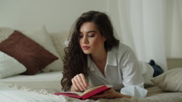 一个年轻的黑头发姑娘躺在床上看书 穿着白衬衫的长发黑发女人在上班前看书 高质量的4K镜头 — 图库视频影像