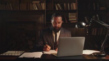 Resmi takım elbiseli bir adam dizüstü bilgisayarın önünde oturur ve belgeleri kontrol eder. Erkek işadamı ofisinde otururken hisse senedi imzalıyor. Yüksek kalite 4k görüntü