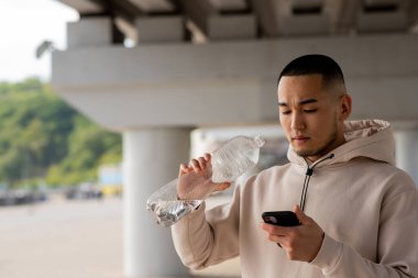 Sıradan kapüşonlu Asyalı bir adam açık hava eğitiminden sonra bir şişe suyla akıllı telefon kullanıyor. Yüksek kalite fotoğraf