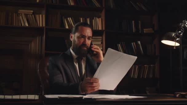 Чоловік Діловому Костюмі Сидить Столом Тримаючи Документи Руці Розмовляючи Телефону — стокове відео