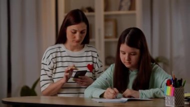 Bir kadın telefonundaki haberlere bakar ve düzenli olarak kızının ev ödevini nasıl yaptığını kontrol eder. Kız masada oturuyor ve bir deftere yazıyor. Annesi de yanında oturuyor.