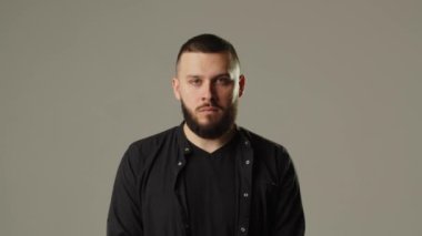 Gri arka planda kameraya poz veren genç sakallı adam. Siyah kazaklı bir adam gri bir duvara yaslanır ve dikkatle kameraya bakar. Yüksek kalite 4k görüntü
