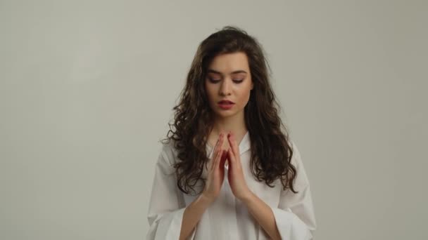 一个穿着白衬衫的漂亮女人站在摄像机前 双手紧握着祈祷 一头长长的黑发站在灰蒙蒙的背景上祈祷着 许下了一个愿望 高质量的4K镜头 — 图库视频影像