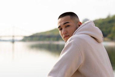 Genç bir adam nehrin yanında duruyor ve omzunun üzerinden kameraya bakıyor. Beyaz kapüşonlu Asyalı bir adam bulanık bir nehrin arka planına karşı kameraya poz veriyor. Yüksek kalite fotoğraf