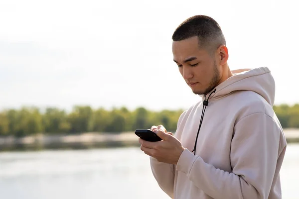 stock image An Asian man stands on the background of the river and taps his finger on the screen of his smartphone. A man in a hoodie watches something on his smartphone while walking along the promenade. High