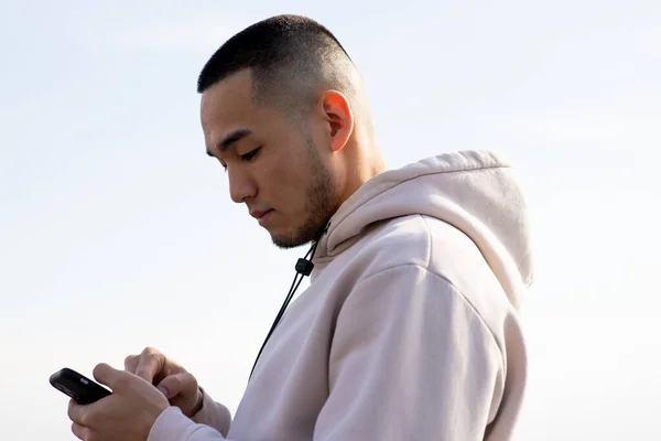 stock image A young man in a hoodie looks at the screen of his smartphone. An Asian guy stands against the background of a cloudy sky and taps on the smartphone screen. High quality photo