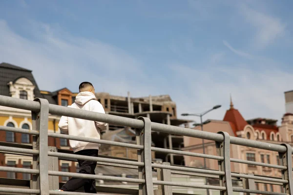 若い男が市内を背景に橋を渡って走っている 白いパーカーを着た男が街中を朝ジョギングしている 高品質の写真 — ストック写真