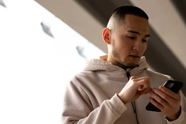 stock image A young man with a short haircut and stubble looks at the screen of his smartphone. An Asian guy in a white hoodie is standing outdoors and looking at something on his phone. High quality photo