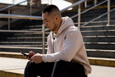 Beyaz kapüşonlu Asyalı bir adam merdivenlerde oturur ve akıllı telefon ekranına bakar. Genç bir adam dışarıda otururken akıllı telefon kullanıyor. Yüksek kalite fotoğraf