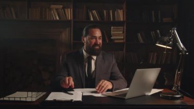 Gülümseyen bir erkek işadamı ofisinde oturur ve laptop ekranına bakar ve sonra bir kağıt parçasına notlar alır. Takım elbiseli sakallı bir adam bir ofiste oturuyor ve belgeleri imzalıyor.