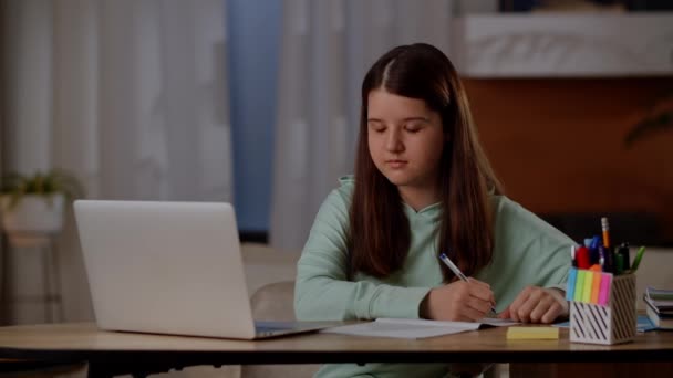 那孩子坐在电脑前的桌子前 在笔记本上记笔记 这个女孩在笔记本电脑上听在线讲座并写摘要 高质量的4K镜头 — 图库视频影像