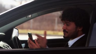 Genç, kıvırcık bir adam arabada oturur ve telefondaki bir şeye bakar. Arabada oturan genç bir adam akıllı telefonun ekranına bakarken bir şeye seviniyor. Yüksek kalite 4k görüntü