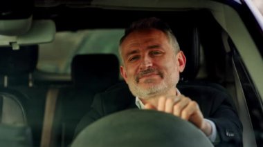 Gülümseyen sakallı bir adam arabada oturuyor ve ellerini direksiyona vuruyor. Bir arabanın direksiyonunda oturmuş kahve içen takım elbiseli gülümseyen bir adamın önden görünüşü.