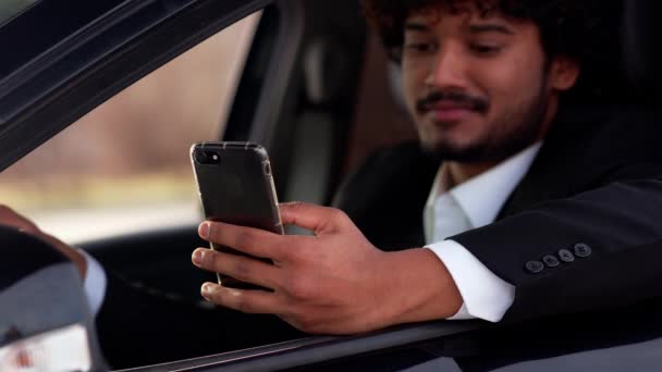 若い巻き毛の男は車の中に座って 彼の電話にメッセージを入力します ビジネススーツの笑顔の男が車のカップからコーヒーを飲み スマートフォンを使用しています 高品質4K映像 — ストック動画