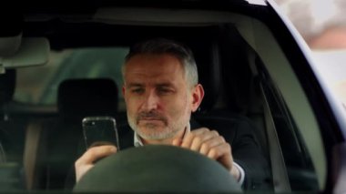 Elinde bir telefonla arabada oturan iş elbisesi giymiş sakallı bir adamın önden çekimi. Direksiyonda oturan ve akıllı telefondan mesaj yazan bir adam. Yüksek kalite 4k görüntü
