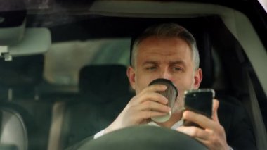 Bir erkek işadamı elinde telefonla arabada oturur ve kahve içer. Takım elbiseli bir adam arabada oturur ve arabası yakıt ikmali yaparken bir fincandan kahve içer. Yüksek kalite 4k görüntü
