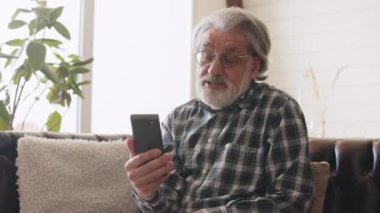Sakallı yaşlı adam video görüşmesi yapıyor. Gözlüklü ve kareli gömlekli büyükbaba telefonda konuşuyor. Yüksek kalite 4k görüntü