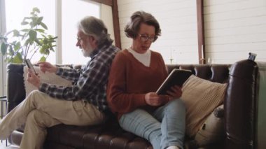 Kanepede otururken akıllı telefon ve dijital tablet kullanan emekli çift. Kıdemli çift alet kullanmayı öğreniyor. Yüksek kalite 4k görüntü