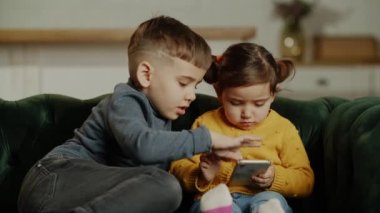 İki küçük çocuk cep telefonunda online ebeveyn kontrol uygulaması kullanıyor, çocuklar için oyun oynuyor, dijital cihazları paylaşıyor, evdeki rahat koltukta dinleniyor. Yüksek kalite 4k görüntü