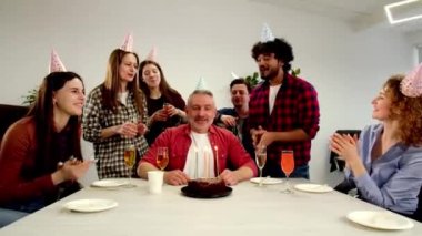 Beyaz saçlı, şenlikli şapkalı bir adam arkadaşlarıyla birlikte oturur ve pastadaki mumları söndürür. Ofis çalışanları iş arkadaşlarının doğum gününü kutluyorlar. Yüksek kalite 4k görüntü